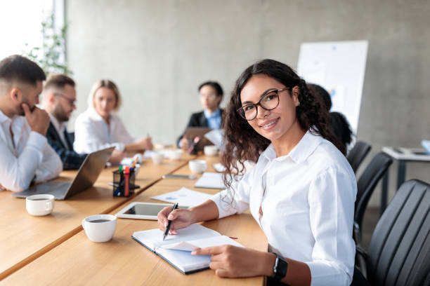 Mexican Businesswoman Posing Taking Notes And Smiling To Camera Sitting At Corporate Business Meeting With Coworkers In Office. Female Entrepreneurship Career Concept